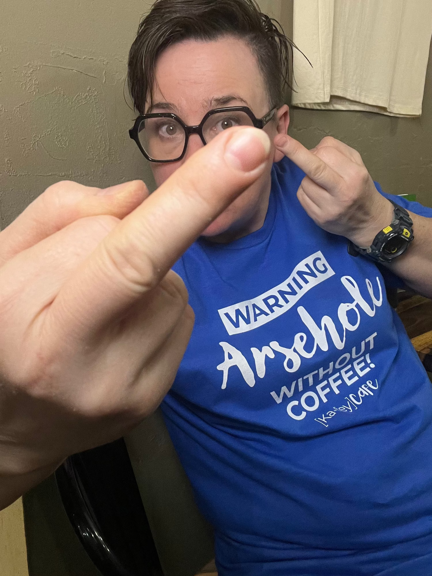 A person wearing a bright blue graphic t-shirt with the text 'WARNING Arsehole WITHOUT COFFEE!' in white, giving the middle finger in an indoor setting.