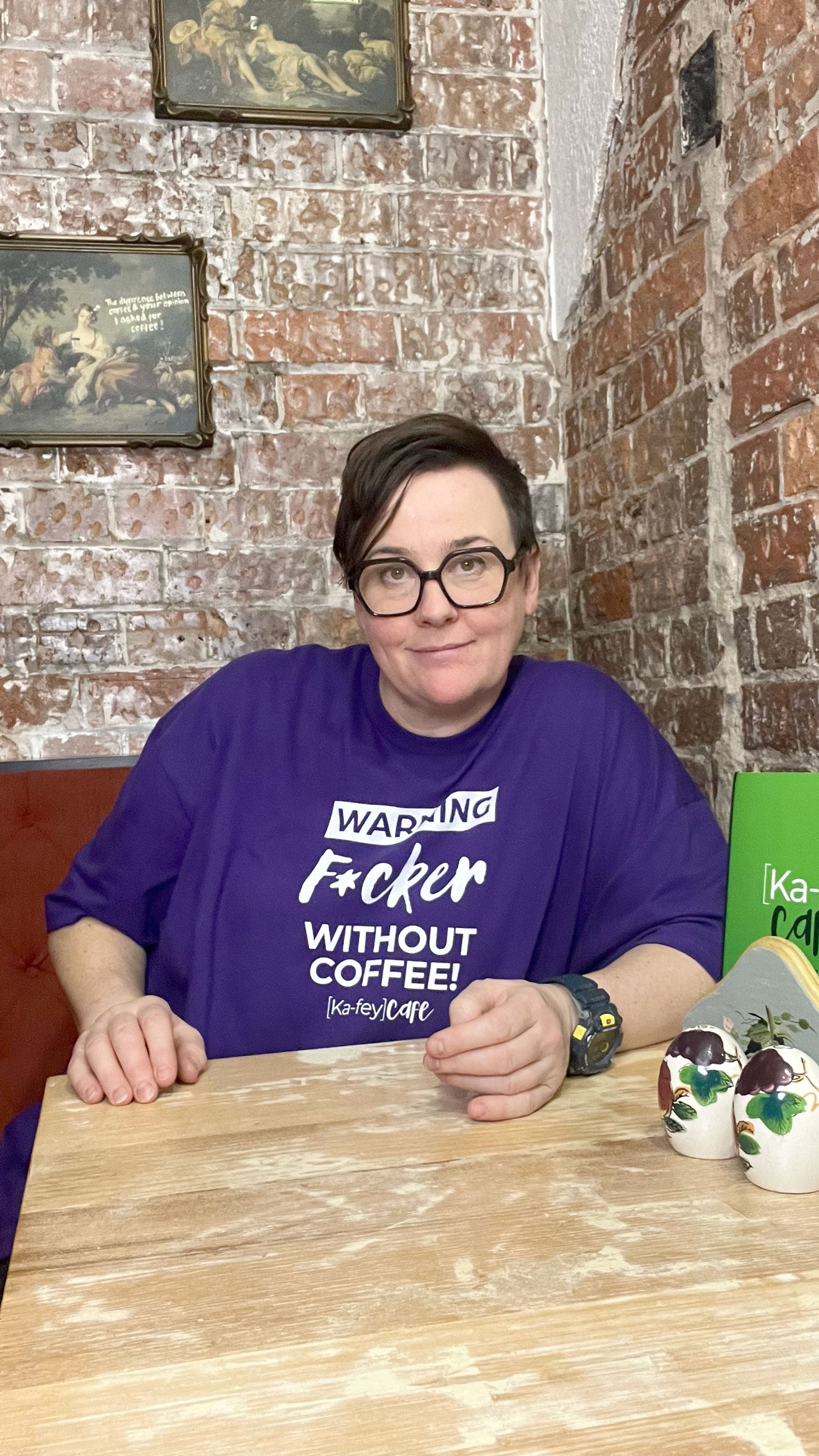 A person wearing a purple graphic t-shirt with white text that reads 'WARNING F*cker WITHOUT COFFEE!  Sat at a wooden table in front of a brick wall.
