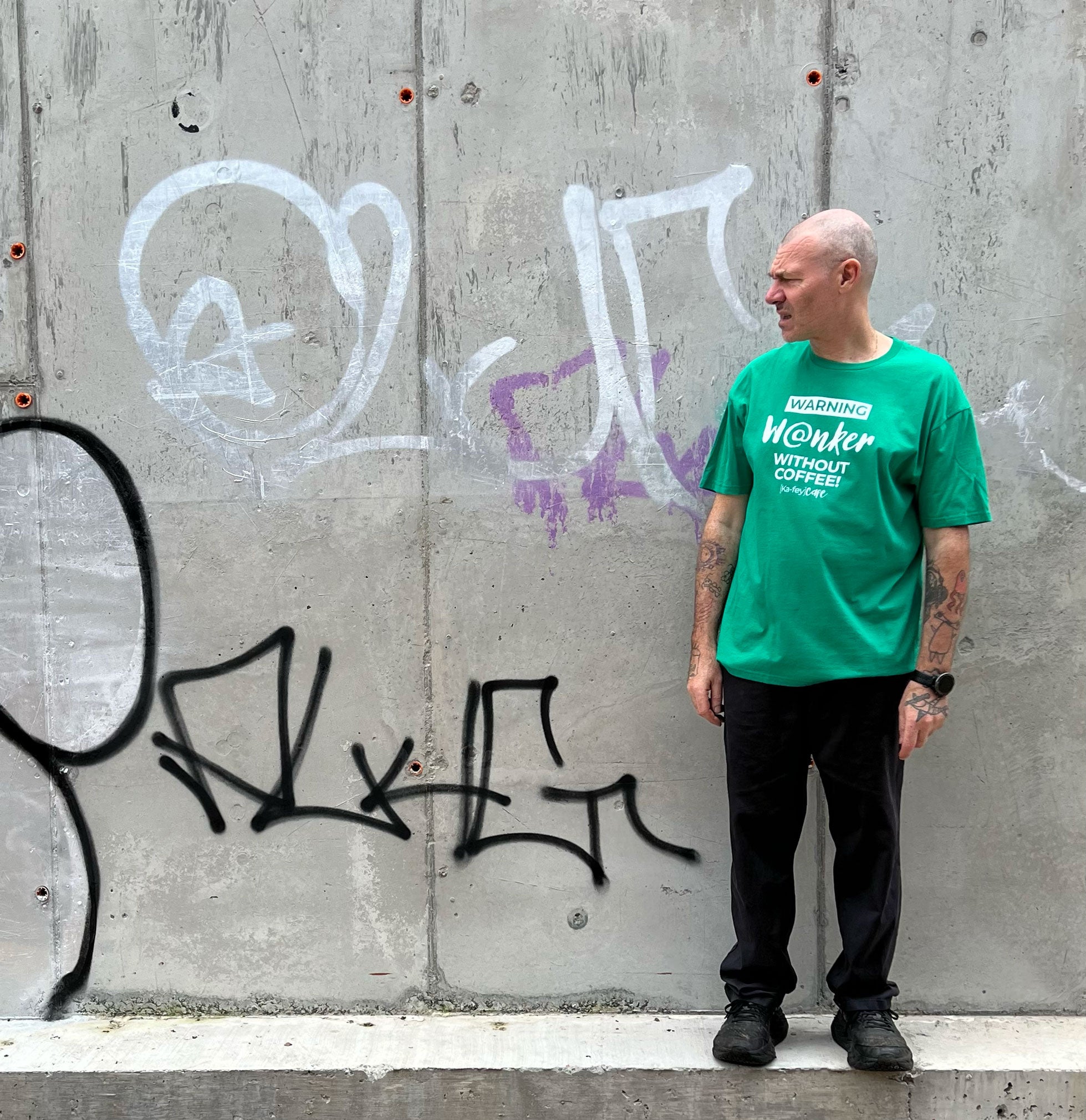 A man with tattoos stands against a concrete wall with graffiti, wearing a bright green t-shirt with white text that reads 'Wanker WITHOUT COFFEE' and black pants.